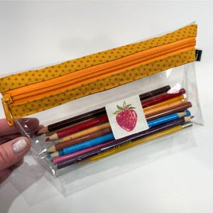 May include: Clear pencil case with a yellow polka dot fabric top and orange zipper. The case contains a variety of colored pencils and a label with a strawberry illustration.