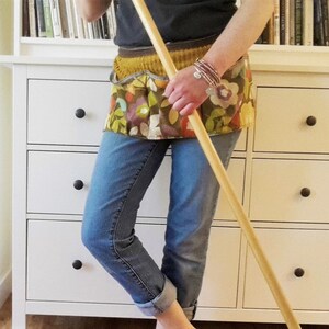 May include: A person wearing blue jeans and a floral apron with a wooden mop in their hand. The person is standing in front of a white dresser with drawers.
