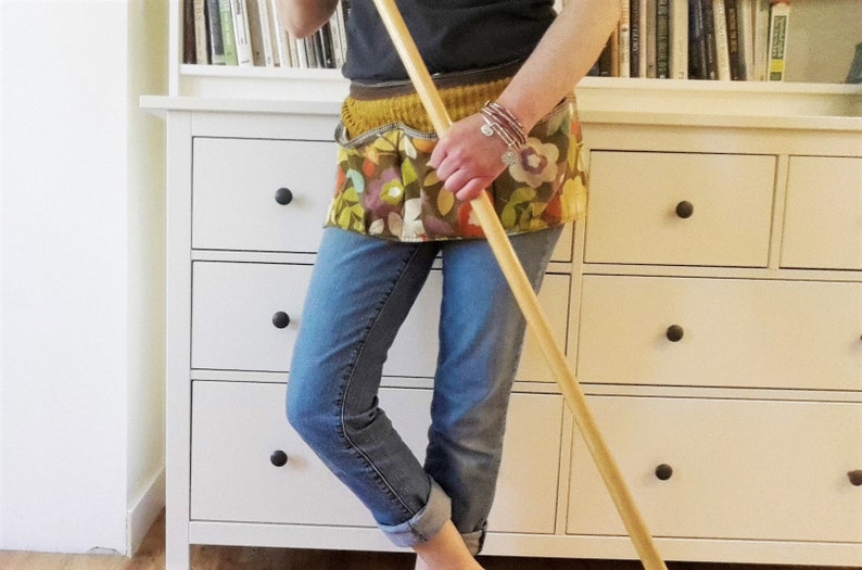 May include: A person wearing blue jeans and a floral apron with a wooden mop in their hand. The person is standing in front of a white dresser with drawers.