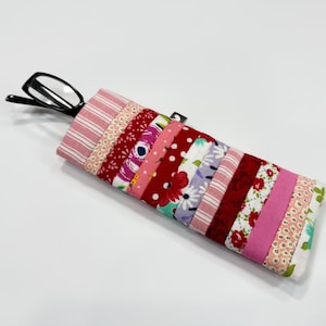 May include: A patchwork glasses case with a variety of fabrics in red, pink, and white. The case has a quilted design with floral and polka dot patterns. Black eyeglasses are partially inserted into the case. The case is handmade.
