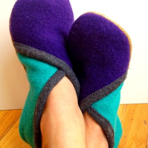 Op de afbeelding: Een paar pantoffels van fleece in paars, turquoise en grijs. De pantoffels hebben een contrasterend kleurendesign met een grijze rand.