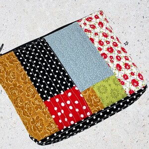 May include: A patchwork fabric zippered pouch with a black zipper. The pouch is made of different fabrics with patterns of polka dots, floral designs, and solid colors. The pouch is quilted and has a black zipper.