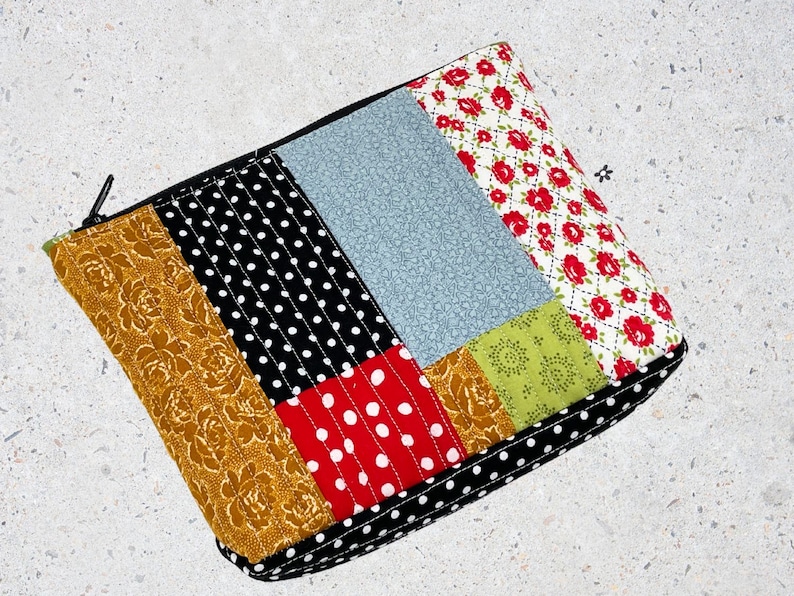 May include: A patchwork fabric zippered pouch with a black zipper. The pouch is made of different fabrics with patterns of polka dots, floral designs, and solid colors. The pouch is quilted and has a black zipper.