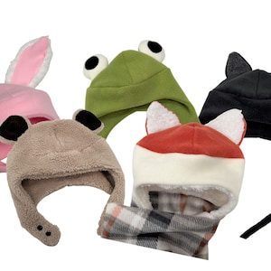 May include: Five different animal-themed fleece hats. The hats are in various colors, including pink, green, brown, black, and red. Each hat has a unique animal feature, such as bunny ears, frog eyes, bear ears, bat ears, and fox ears.