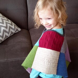 May include: A young child wearing a colorful patchwork poncho made from different types of knit fabric. The poncho has a red, white, green, blue, and purple color scheme.
