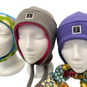 May include: Three fleece hats with ear flaps and a tie string. The hats are gray, purple, and blue and green with white polka dots. Each hat has a small black square patch with a white letter "J" on it.