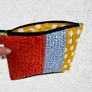 May include: A colourful zippered pouch with a patchwork design. The pouch is made of four different fabrics: red with a floral pattern, blue with white polka dots, yellow with white polka dots, and a solid green fabric.
