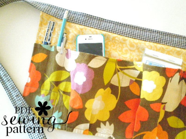 May include: A brown fabric apron with multiple pockets, featuring a floral print in shades of yellow, orange, and purple. The apron has a black and white checkered trim and includes a cell phone pocket, a pencil holder, and a pocket for a small notepad.