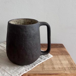May include: A dark, textured ceramic mug with a rounded body and a rectangular handle. The interior of the mug has a lighter, speckled finish. It sits on a white cloth atop a wooden surface, with a plain white wall in the background.