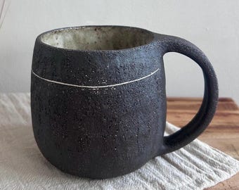 Op de afbeelding: Een zwarte keramische mok met een textuur en een witte horizontale lijn. De binnenkant is lichter van kleur. De mok heeft een groot oor en staat op een witte doek op een houten oppervlak.