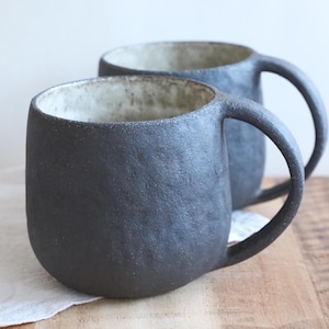 Peut inclure: Deux tasses en céramique artisanale présentant un extérieur noir anthracite texturé et un intérieur contrastant blanc cassé moucheté. Chaque tasse possède une forme arrondie et organique avec une anse incurvée et robuste sur le côté.