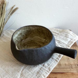 Pode incluir: Tigela de cerâmica preta feita à mão com uma alça. O interior tem um esmalte salpicado verde e castanho terroso. A tigela está sobre um pano creme, com ervas secas ao fundo. Ideal para servir sopas ou ensopados.