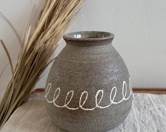 May include: A speckled gray ceramic vase with a white spiral design. The vase has a rounded body and a narrow neck. Dried grasses are to the left of the vase, and the vase sits on a white cloth.
