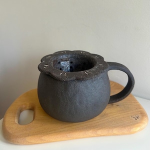 May include: A dark grey ceramic tea cup with a floral rim and a handle. The cup is placed on a light wooden chopping board. The cup has a textured surface and a dark interior.