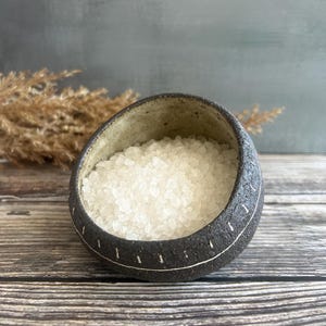 Puede incluir: Un pequeño cuenco de cerámica gris oscuro lleno de sal gruesa blanca. El cuenco tiene forma redondeada con un exterior texturizado y un diseño cosido blanco alrededor del borde. El fondo es una mezcla borrosa de gris y marrón.