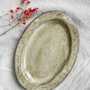 Könnte beinhalten: Eine ovale Servierplatte aus Keramik mit gesprenkelter, cremefarbener Glasur und dunklem Rand. Die Platte wird auf einer weißen, strukturierten Oberfläche präsentiert, mit einem Zweig roter Blumen in der oberen linken Ecke.