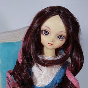 May include: A doll with long, curly, dark brown hair with pink streaks. The doll is wearing a blue dress with white lace trim and a white lace top. The doll has blue eyes and a light complexion.