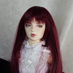 Puede incluir: Una muñeca con el pelo largo, liso y rojo burdeos. La muñeca lleva un vestido blanco con encaje.