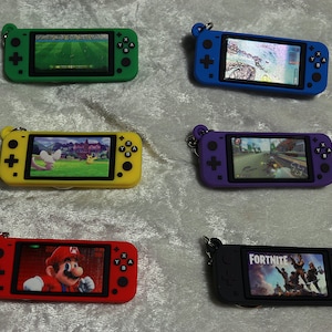 May include: Six colorful keychain replicas of the Nintendo Switch handheld gaming console. Each keychain features a different game screen, including soccer, Pokemon, racing, Mario, and Fortnite.