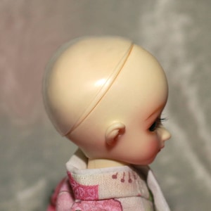 May include: A doll head with a smooth, light-colored skin tone and a clear plastic dome covering the top of the head. The doll has a closed mouth and a slightly parted eyelid. The doll is wearing a white and pink patterned dress.