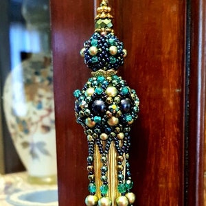 Può includere: Un nappa di perline con catena e chiusura dorate. La nappa presenta un gruppo di perline blu scuro, verde acqua e dorate, con una frangia di filo dorato. L'accento decorativo è appeso su uno sfondo di legno scuro.