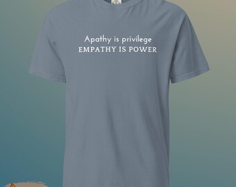 May include: A dusty blue t-shirt with the text "Apathy is privilege, EMPATHY IS POWER" in white. The shirt has short sleeves and a crew neck. The shirt is made of a soft, comfortable material.