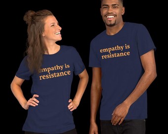 May include: Two people wearing navy blue t-shirts with the text "empathy is resistance" in a gold color. The t-shirts are short-sleeved and appear to be made of a soft material. The background is black.