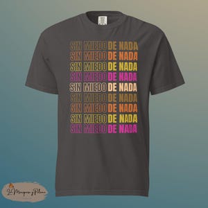 May include: Dark grey t-shirt featuring the phrase "SIN MIEDO DE NADA" in a repeating pattern. The text is in shades of brown, yellow, and pink. The t-shirt has short sleeves and a crew neck.
