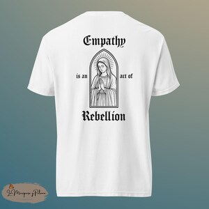 May include: White t-shirt with a graphic on the back featuring a black and white illustration of a religious figure within an arched frame. The text reads "Empathy is an act of Rebellion."