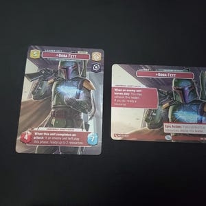 May include: Two Star Wars trading cards featuring Boba Fett. The cards have a purple and gray color scheme with text and illustrations of the character. One card has the text "When this unit completes an attack: If an enemy unit left play this phase, ready up to 2 resources."
