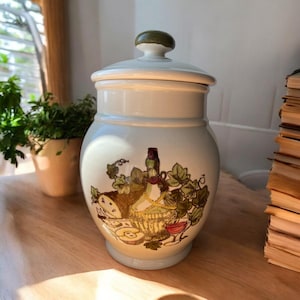 May include: A white ceramic jar with a lid, featuring a hand-painted design of a bottle of wine, grapes, and cheese in a basket. The jar has a light blue glaze and a green lid.