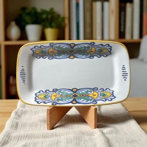 May include: Rectangular ceramic serving platter with a white base, adorned with ornate blue, green, and yellow floral designs. The platter has a yellow border and is displayed on a wooden stand, suitable for serving food.