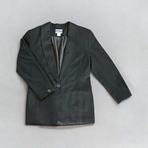 Può includere: Un blazer nero con un unico bottone di chiusura e un colletto largo. Il blazer ha una tasca sul lato sinistro e un'etichetta che recita "P.S. from Aeropostale".