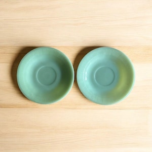Puede incluir: Dos platillos vintage de vidrio verde claro con diseño acanalado. Los platillos son redondos con una hendidura central para una taza. El color es un verde suave y apagado, sobre una superficie de madera clara.