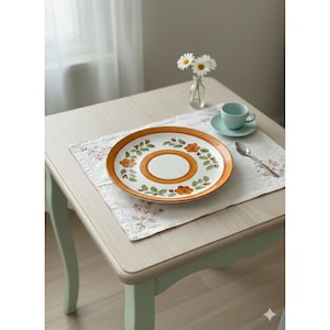 May include: A vintage-style ceramic plate with a brown border and floral design, set on a floral embroidered placemat. A small blue teacup and saucer, along with a spoon, are on the table. A small vase of daisies sits nearby.
