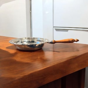 May include: A silver serving dish with a wooden handle. The dish has a decorative rim and is sitting on a wooden table.
