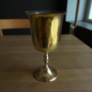 Puede incluir: Un cáliz dorado con un cuenco ancho y redondeado y un tallo delgado. El cáliz tiene un acabado pulido, que refleja la luz. La base es circular y ligeramente ensanchada. El cáliz probablemente está hecho de latón o un metal similar.