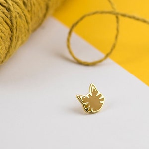 May include: A gold-colored cat face enamel pin with a brown striped design. The pin is set against a white and yellow background, with a spool of yellow thread in the upper left corner.