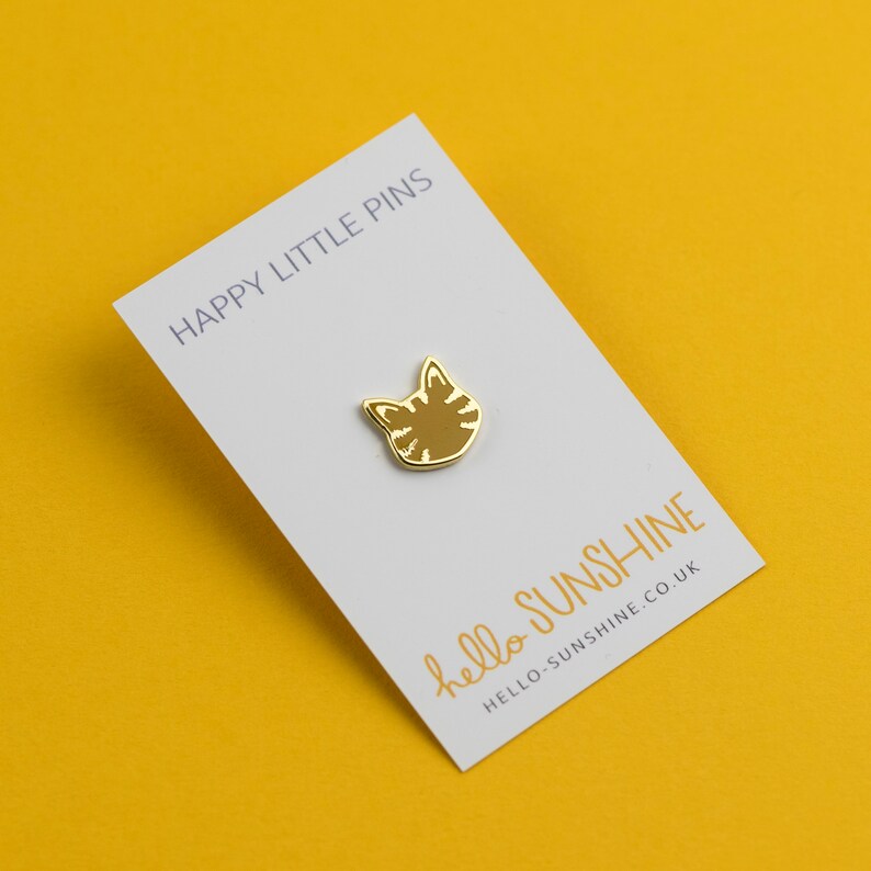 May include: A gold-colored cat face pin with striped details, displayed on a white card. The card has the text "HAPPY LITTLE PINS" at the top and "hello sunshine" with a website address at the bottom. The background is a solid yellow.