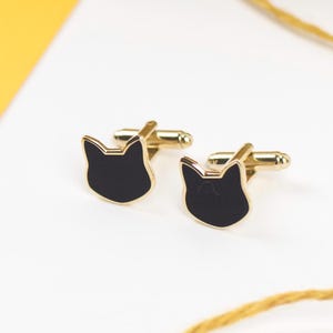 May include: A pair of gold-toned cufflinks featuring a black cat face design. The cat faces have a gold outline and are set against a white background. The cufflinks are a stylish accessory.