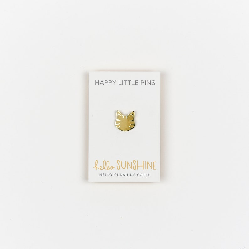 May include: A gold-colored cat face pin on a white card. The card reads "HAPPY LITTLE PINS" at the top and "hello SUNSHINE" with a website address at the bottom. The pin is centered on the card.