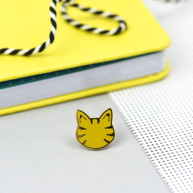 May include: A yellow cat face enamel pin with black stripes and a gold outline. The pin is set on a light gray surface, with a yellow notebook and a black and white cord in the background.
