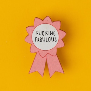 Könnte beinhalten: Ein rosa Holz-Auszeichnungsband mit den Worten "FUCKING FABULOUS" in schwarzem Text auf einem weißen Kreis. Das Band hat einen gewellten Rand und zwei Bandenden. Der Hintergrund ist einfarbig gelb.