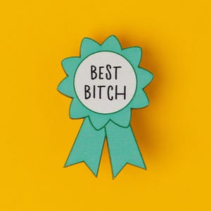 Könnte beinhalten: Eine helltürkisfarbene Schleife mit den schwarzen Worten "BEST BITCH" auf einem weißen Kreis. Die Schleife befindet sich auf einem leuchtend gelben Hintergrund.