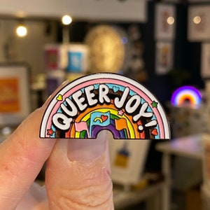Könnte beinhalten: Eine regenbogenförmige Emaille-Anstecknadel mit den Worten "Queer Joy!" in weißen Buchstaben. Der Regenbogen hat einen rosa, orange, gelben, grünen, blauen und violetten Streifen. Um den Regenbogen herum befinden sich kleine Sterne und Herzen.