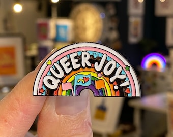 May include: A rainbow-shaped enamel pin with the words "Queer Joy!" in white letters. The rainbow has a pink, orange, yellow, green, blue, and purple stripe. There are small stars and hearts around the rainbow.