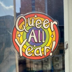 May include: A rainbow-colored sticker with the text "Queer All Year!" in a retro font. The sticker has a white background and is surrounded by a rainbow-colored border.