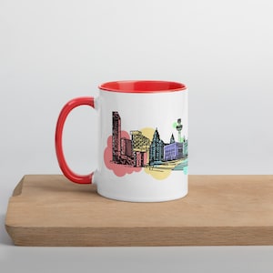 Könnte beinhalten: Weißer Keramikbecher mit roter Innenseite und Henkel. Der Becher zeigt eine farbenfrohe Strichzeichnung einer Stadtlandschaft, mit Gebäuden und Wahrzeichen in Schwarz, akzentuiert durch rote, gelbe und blaue Farbtöne. Der Becher steht auf einer Holzoberfläche.