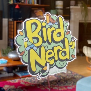 May include: A sticker featuring the text "Bird Nerd" in yellow with a black outline, set against a green backdrop. The design includes illustrations of birds and leaves. The artist's signature, "Sophie Green," is visible at the bottom.