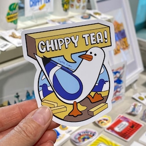 May include: A white seagull with blue wings and orange feet stands on a yellow brick path. The seagull is holding a speech bubble that says "Chippy Tea!" in black text. The background is a blue sky with white clouds. The sticker is signed "Sophie Green".